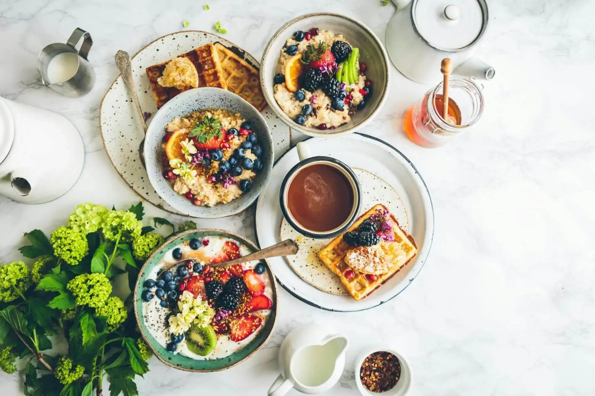 Un desayuno luminoso y acogedor sobre una mesa de mármol, con coloridos cuencos de avena cubiertos de frutas y semillas, gofres, té y un tarro de miel.