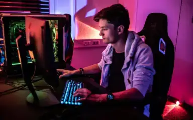 Un joven con capucha utiliza atentamente un ordenador en una habitación poco iluminada con vibrantes luces de neón. La escena destila un ambiente de juego concentrado y moderno.