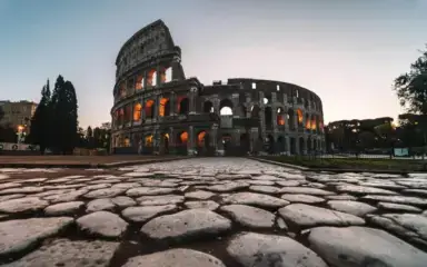 Vista en ángulo bajo del Coliseo de Roma al atardecer, con sus arcos iluminados con luces cálidas. El primer plano de adoquines añade profundidad histórica. Ambiente sereno.