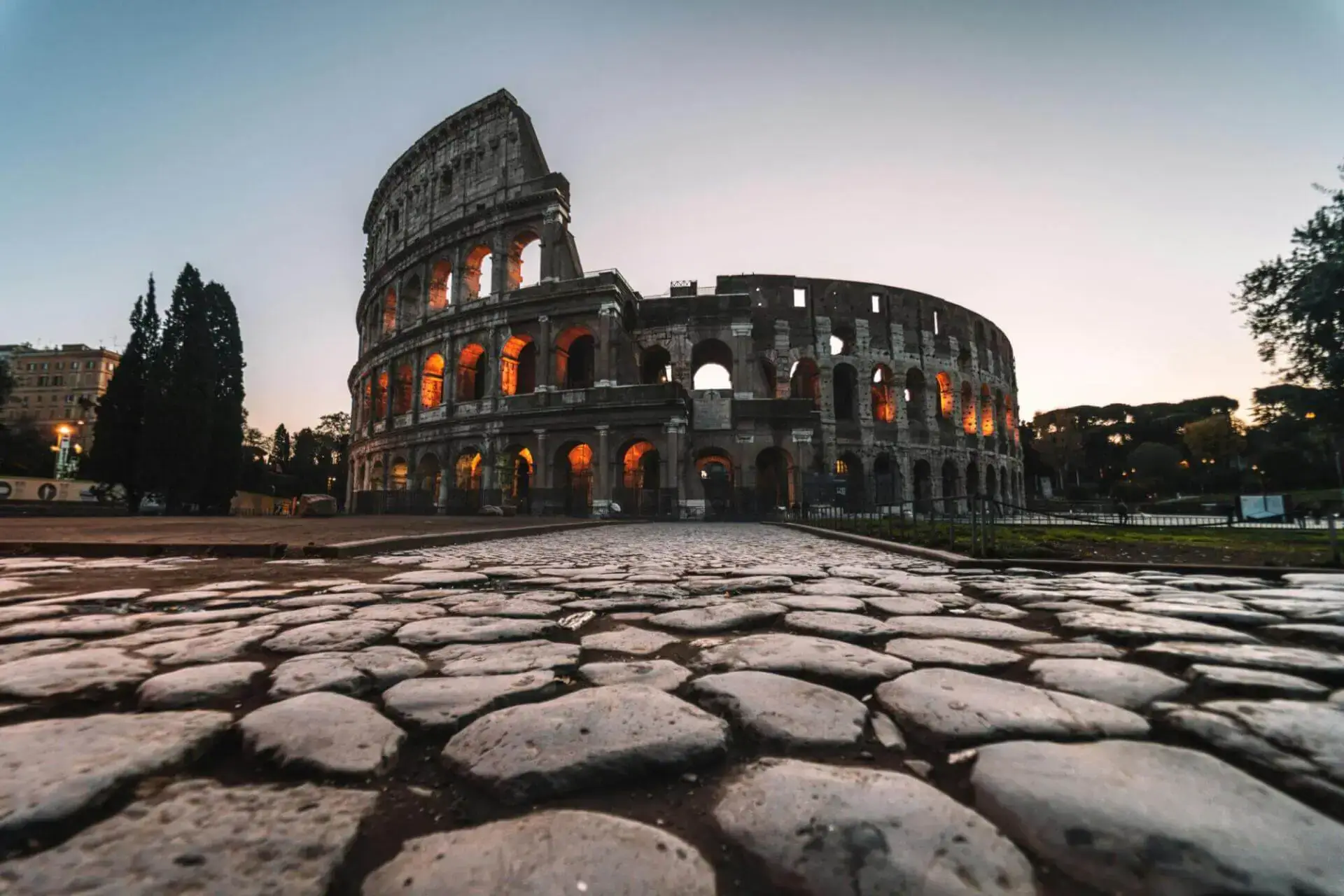 Vista en ángulo bajo del Coliseo de Roma al atardecer, con sus arcos iluminados con luces cálidas. El primer plano de adoquines añade profundidad histórica. Ambiente sereno.