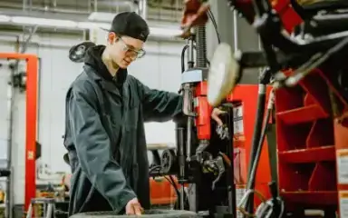 Un joven mecánico con gorra y gafas de seguridad maneja una máquina junto a un neumático en un garaje. El ambiente es laborioso y concentrado.