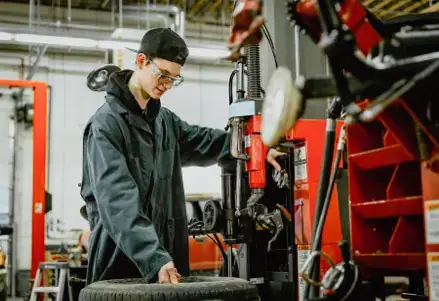 Un joven mecánico con gorra y gafas de seguridad maneja una máquina junto a un neumático en un garaje. El ambiente es laborioso y concentrado.