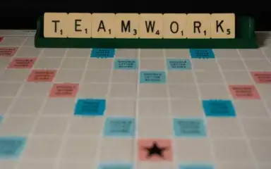 Las fichas de Scrabble deletrean "TRABAJO EN EQUIPO" en un estante sobre un tablero de juego. El tablero presenta casillas de bonificación en azul y rosa, transmitiendo un tema de colaboración.