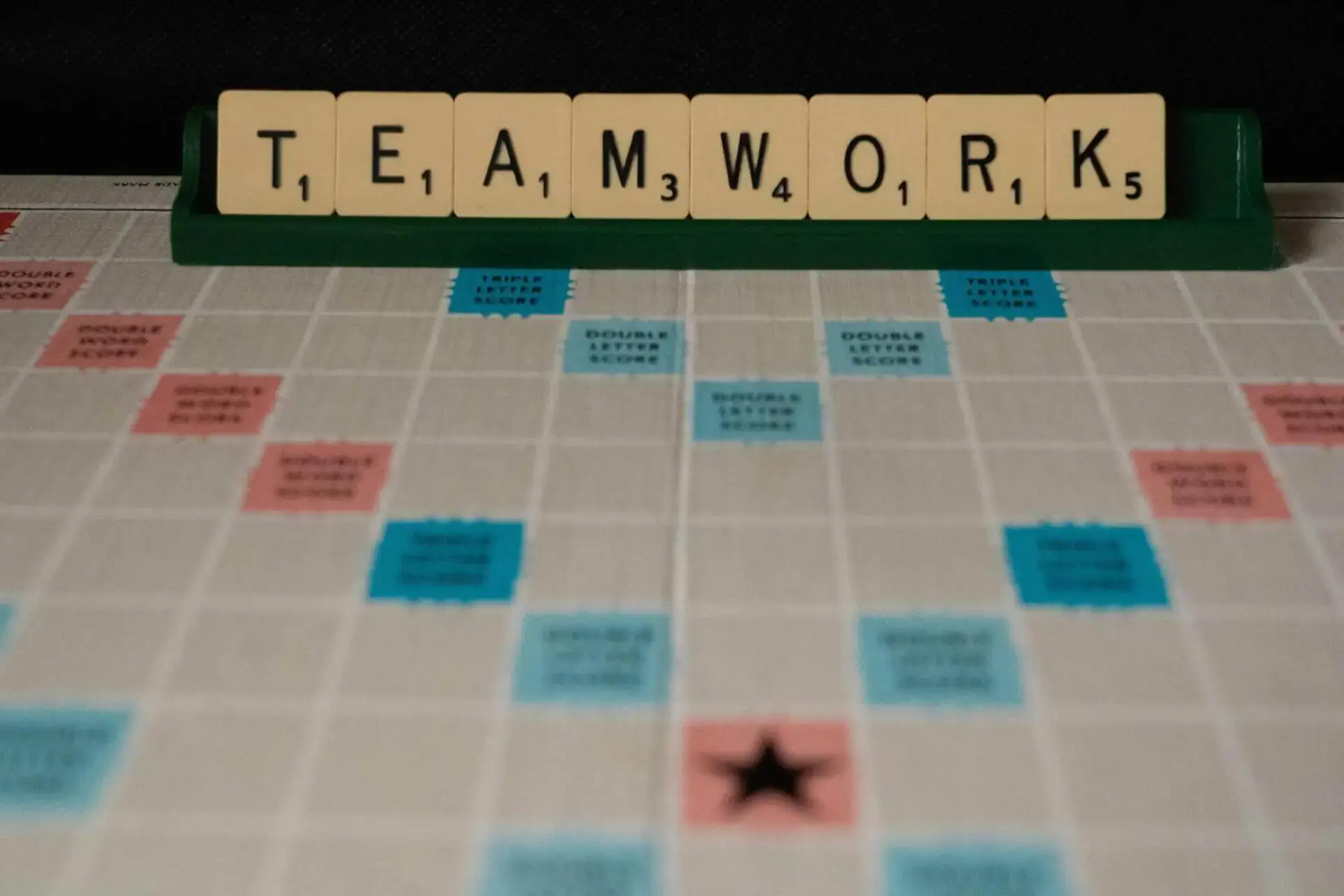 Las fichas de Scrabble deletrean "TRABAJO EN EQUIPO" en un estante sobre un tablero de juego. El tablero presenta casillas de bonificación en azul y rosa, transmitiendo un tema de colaboración.