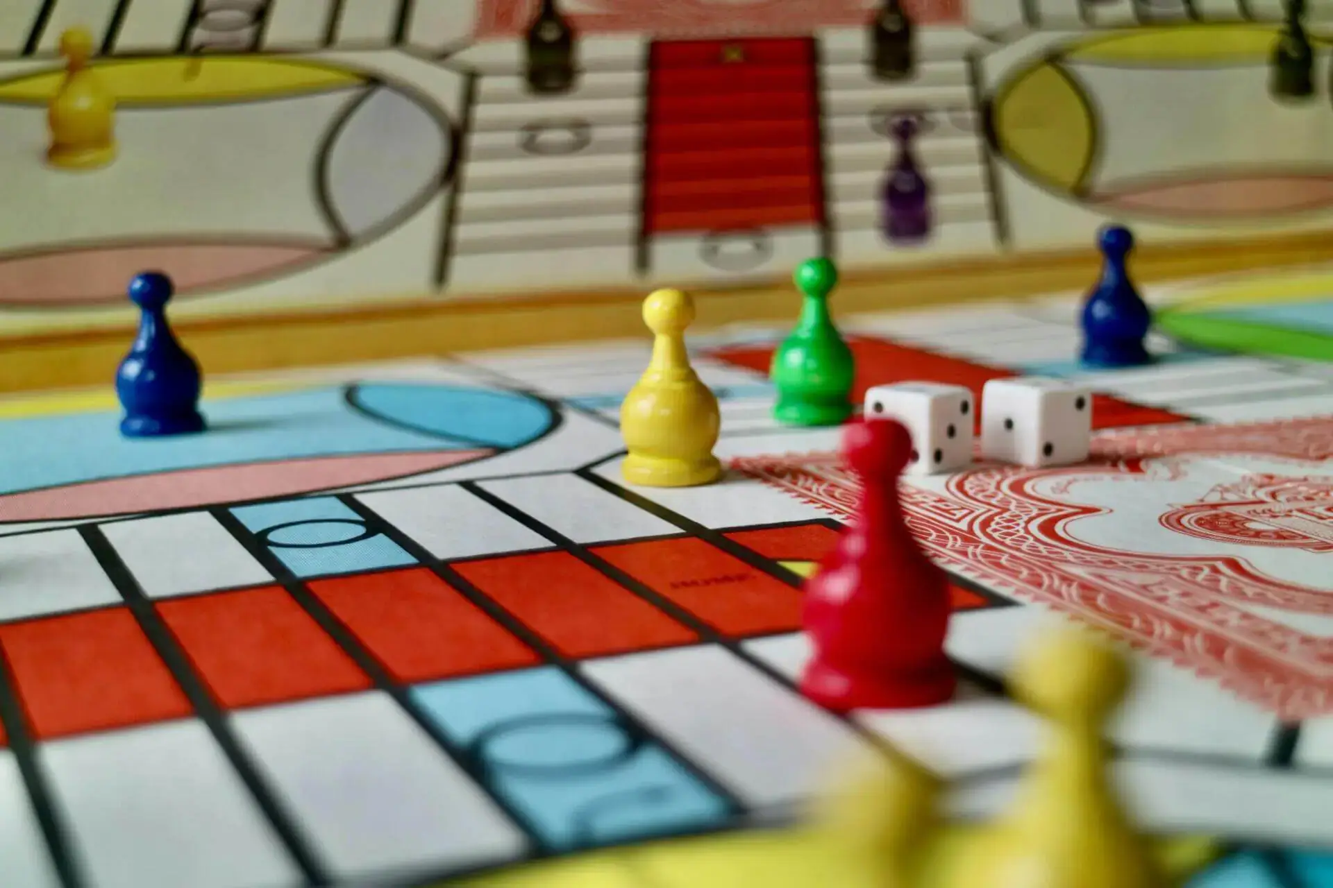 Colorido tablero de juego con peones amarillos, rojos, azules y verdes dispuestos cerca de dos dados blancos. El fondo muestra patrones intrincados, lo que sugiere un juego animado y estratégico.