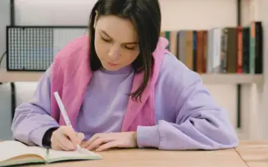 Una mujer con sudadera lavanda y bufanda rosa escribe en un cuaderno en una mesa de madera. Una estantería con libros aparece borrosa al fondo.
