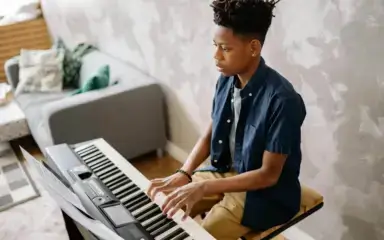 Adolescente tocando un teclado en una acogedora habitación con una pared estampada y un sofá. La expresión concentrada sugiere un ambiente serio pero tranquilo.