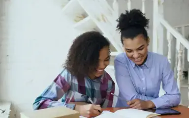 Dos mujeres sentadas a una mesa, sonríen mientras se entretienen con un cuaderno abierto. El acogedor fondo blanco y la luz natural añaden un ambiente cálido y alegre.